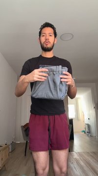 Άνδρας με σγουρά μαλλιά και γένια στέκεται μέσα, κρατώντας ένα ζευγάρι διπλωμένα γκρι παντελόνια, φοράει μαύρο πουκάμισο και μπορντό σορτσάκι.