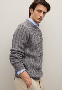 Grauer Strickpullover mit Zopfmuster und rundem Ausschnitt, getragen über einem hellblauen Hemd mit Kragen. Die Struktur ist grob, mit sichtbaren Mustern und gerippten Bündchen.
