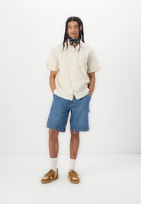 Camisa de botón de manga corta color crema con diseño bordado, combinada con pantalones cortos de mezclilla azules. Zapatillas marrones con acentos texturizados y calcetines blancos.