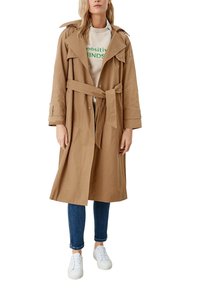 Beige trench coat with a belted waist, large collar, and snap buttons, worn with a grey sweatshirt, blue jeans, and white sneakers.