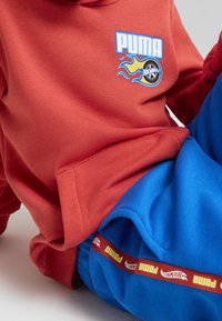Ein Kind tr�ägt einen roten Hoodie mit Puma- und Hot-Wheels-Graphic-Design sowie eine blaue Sporthose mit Streifen in Puma- und Hot-Wheels-Logo-Details und sitzt auf einer weißen Oberfläche.