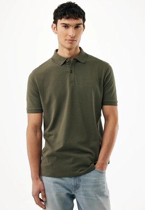 Junger Mann, der ein olivgrünes Poloshirt mit kurzen Ärmeln und hellblaue Jeans trägt, mit der Hand in der Tasche, vor einem weißen Hintergrund.