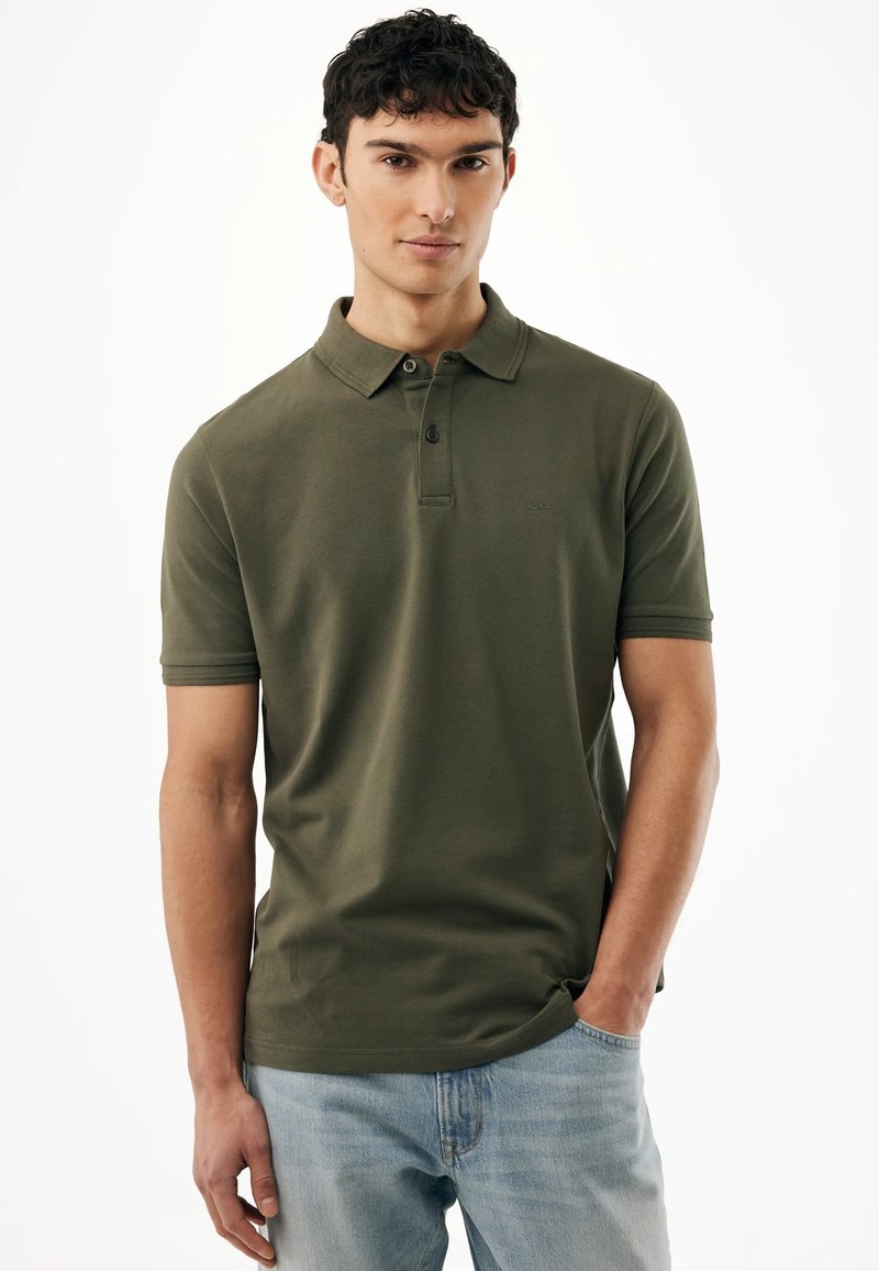 Joven vestido con una camisa polo de manga corta verde oliva y vaqueros azul claro, con la mano en el bolsillo, frente a un fondo blanco.