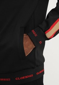 Ruka v kapse černé bundy; bunda má červené a béžové pruhy na rukávech, černé žebrované manžety a lem s červeným nápisem "GLORIOUS".