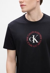 Czarny bawełniany t-shirt z okrągłym logo z białym i czerwonym tekstem, w tym "Calvin Klein" i "Since 1968" wokół centralnego wzoru "CK".