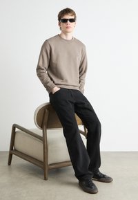 Sudadera marrón, jeans negros, gafas de sol y zapatillas oscuras. Modelo sentado en una silla tapizada clara con patas de madera.