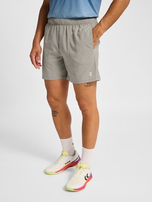 Mann trägt hellgraue Shorts mit elastischem Bund, weiße Socken und weiße Sneakers mit gelben, pinken und violetten Akzenten und steht auf einem weißen Boden.