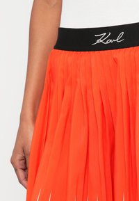 Redőzött narancssárga szoknya fekete derékrésszel, fehér "Karl" hímezéssel, könnyű anyagból, sima textúrával.