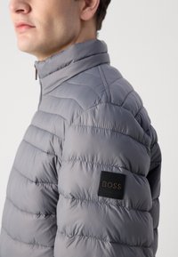 Gray puffer jacket with a high collar and horizontal quilted texture. Features a branded black patch on the left shoulder.