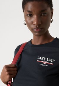 Juodas marškinėliai su "GANT 1949" logotipu balta ir raudona spalva; trumpomis rankovėmis, apvaliu kaklu; modelis dėvi raudoną maišelio diržą ir sidabrines auskarus.