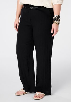 Persoon die losse zwarte broek draagt met een dunne zwarte riem, een panterprint sjaal om de pols, gouden armbanden en witte sandalen versierd met schelpen.