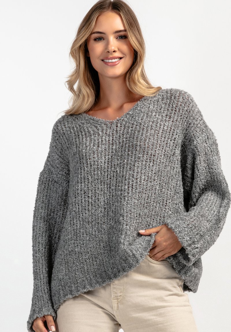 Szary sweter dziewiarski o luźnym kroju, z dekoltem w serek i prążkowaną fakturą. Posiada boczne rozcięcia i długie rękawy, noszony na beżowych spodniach.