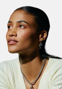 Modelka s tmavými vlasmi, ktorá má na sebe strieborný náhrdelník a visiace náušnice. Top je svetložltý, pruhovaný a má výstrih do V.