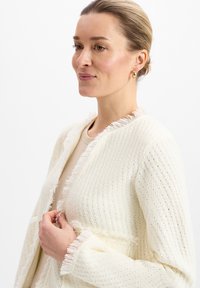 Cardigan leggero in crema con bordi frangiati, motivo a maglia testurizzata, aperto sul davanti e maniche lunghe, indossato su una maglietta beige.