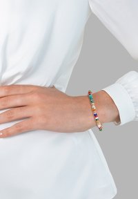 Farbiger Perlenarmband mit verschiedenen Formen und Größen von Perlen in Grün, Rot, Blau, Weiß und Gold, getragen an einem Handgelenk mit einem weißen Hemd.
