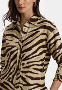 Beige skjorte med mørkebrune tigerstriper, med krage, flere knapper foran og opprullede ermer. Teksturet stoff.