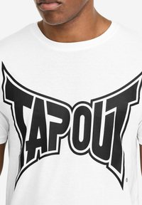 Weißes Baumwoll-T-Shirt mit einem großen schwarzen "TAPOUT"-Logo mit scharfen Kanten. Das Design umfasst dicke Umrisse und einen kräftigen, grafischen Stil.