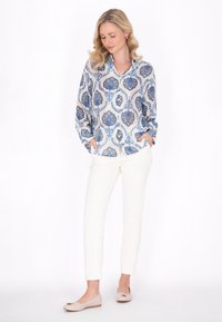 Locker sitzende, langärmelige Bluse in Weiß mit blauen Blumenmustern, kombiniert mit schlanken weißen Hosen und beigen Ballerinas mit dekorativem Akzent.