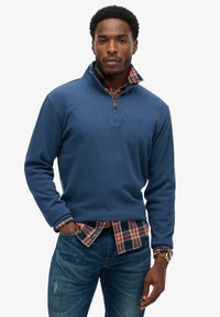 Granatowy pulower typu quarter-zip z teksturowanej tkaniny, z małym logo, nałożony na czerwono-czarną koszulę w kratę, w zestawieniu z ciemnymi dżinsami.