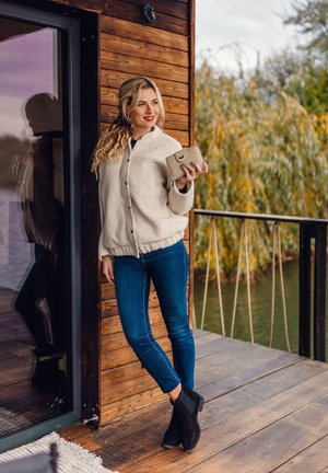 Light beige fleece jacket with buttons, blue skinny jeans, and black ankle boots. Standing on wooden deck, holding a beige wallet.