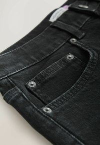 Gros plan d'un jean en denim noir montrant la poche avant avec des rivets métalliques et des passants de ceinture sur un fond clair.
