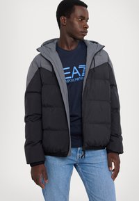 Giacca a vento imbottita con corpo nero e spalle grigie, dotata di cappuccio, chiusura con zip e texture trapuntata. Indossata sopra una t-shirt blu navy con logo.