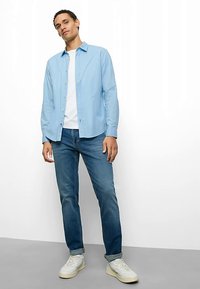 Hellblaues Hemd über einem weißen T-Shirt, kombiniert mit dunkelblauen Jeans und weißen Sneakers. Model steht vor einem neutralen Hintergrund.