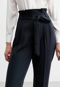Hogewaisted, donkerblauwe broeken met een vastgebonden stoffen riem, geplooide voorkant, taps toelopende benen en een gladde textuur. Witte blouse mouwen zichtbaar.