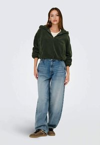 Jovem mulher de pé, vestindo um pullover de fleece verde escuro, jeans de perna larga azul-claro e ténis com estampado de leopardo, contra um fundo simples.