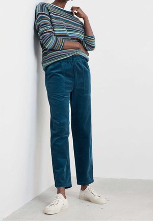 Personne portant un pantalon en velours côtelé bleu sarcelle, un pull rayé à manches longues et des baskets blanches, debout contre un mur et un sol blancs.