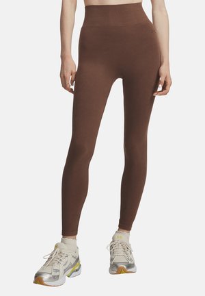 Femme portant des leggings côtelés marron taille haute et des baskets blanches avec des touches jaunes, debout devant un fond clair uni.