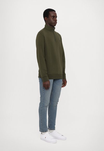 Oliven farvet zip-up sweatshirt med høj krave, ribstrikkede manchetter og søm; parret med lyseblå slim-fit jeans og hvide sneakers.