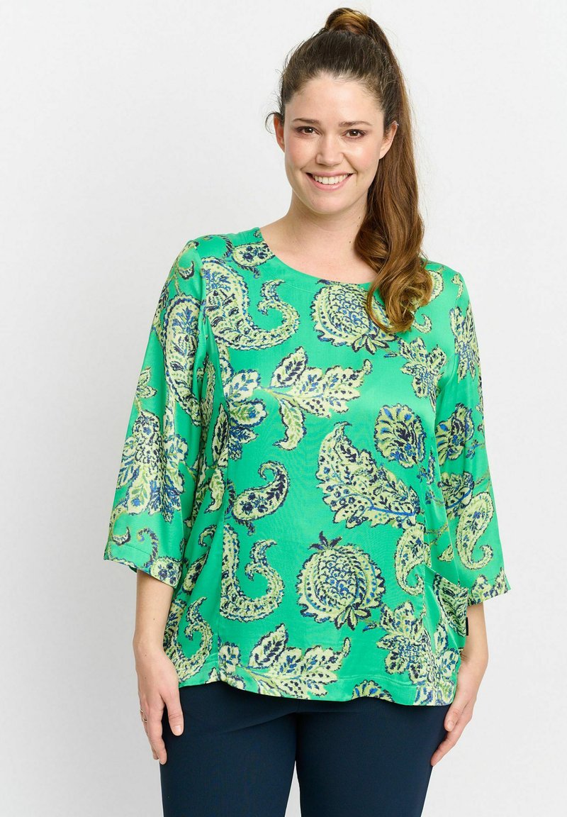 Lächelnde Frau mit langen braunen Haaren im Pferdeschwanz, die eine grüne Bluse mit Blumenmuster und Dreiviertelärmeln sowie dunkle Hosen trägt.