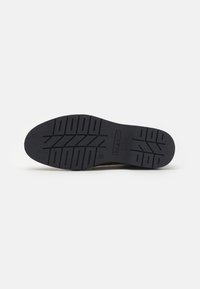 Brown sneaker outsole with a textured black rubber surface. Features a herringbone tread pattern and embossed brand name "bugatti."