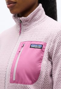 Γυναίκα που φοράει υφασμάτινο fleece μπουφάν με φερμουάρ σε λεβαντί απόχρωση, με φωτεινή ροζ τσέπη στο στήθος που κλείνει με φερμουάρ και ετικέτα με το λογότυπο της Patagonia.