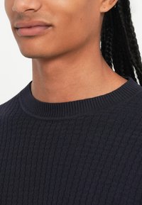 Pulover tricotat bleu marin cu textură ribbed, cu guler rotund și finisaj neted. Fără modele sau accesorii notabile.