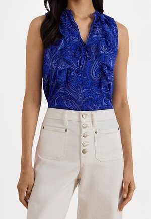 Blouse bleue sans manches à motif cachemire avec un col volanté, associée à un pantalon blanc taille haute avec cinq boutons sur le devant et des poches avant.