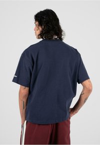 Tamsiai mėlynas medvilninis marškinėliai su trumpomis rankovėmis ir laisvu siluetu. Yra apvalus iškirpimas ir subtilus logotipas ant kairės rankovės.