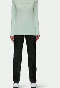 Egy nő világoszöld, hosszú ujjú pólót visel, rajta a "Mammut" logóval, fekete nadrágban és fehér cipőben áll, egy sima fehér háttér előtt.