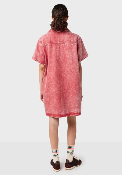 Persoon met de rug naar de camera, gekleed in een rood vervaagd denim jurkje met korte mouwen, witte sokken met regenboogstrepen en donkerbruine sneakers.