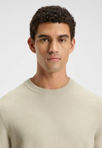 Hellbeiges Sweatshirt mit geripptem Rundhalsausschnitt und einem strukturierten Strickmuster. Das Model zeigt einen neutralen Gesichtsausdruck mit kurzem, lockigem Haar.
