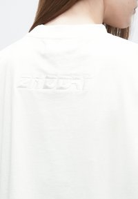 Biele tričko s okrúhlym výstrihom, na chrbte s textúrovaným vyšívaným logom "2NDDAY". Hladká bavlnená tkanina, jednoduchý dizajn.