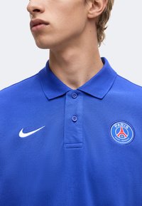 Νέος/Νέα που φοράει μπλε πόλο της Nike με το λογότυπο της Paris Saint-Germain, κοιτάζοντας πλαγίως σε απλό ανοιχτό φόντο.