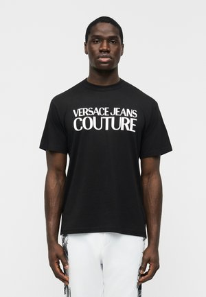 Άνδρας που φοράει μαύρο T-shirt με τη φράση «Versace Jeans Couture» σε λευκά γράμματα, συνδυασμένο με άσπρο παντελόνι που φέρει μαύρα πλευρικά σχέδια.