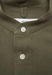 Olivgrünes Leinenhemd mit einem Henley-Kragen, kurzem Knopfverschluss und weißen Vierlochknoopfen. Strukturierter Stoff mit matter Oberfläche.