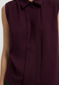 Blusa senza maniche bordeaux con colletto bottonato, caratterizzata da pince verticali sul davanti e un tessuto morbido e leggero.