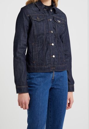 Person iført mørkeblå denimjakke med metal knapper og brystlommer, kombineret med mellemblå denimjeans mod en ensfarvet baggrund.
