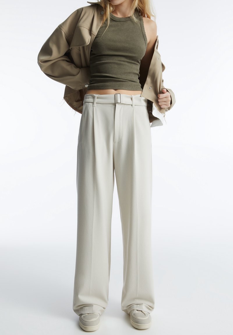 Beige oversize bunda, olivová žebrovaná tílko a smetanové široké kalhoty s opaskem v pase. Outfit doplňují ležérní boty.