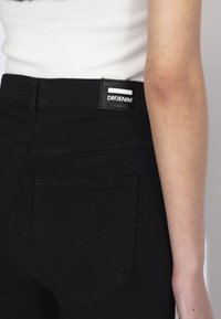Zbliżenie na osobę noszącą czarne dżinsy denim z widoczną tylną kieszenią i markową naszywką z napisem DRDENIM, w zestawieniu z białym prążkowanym topem.