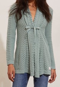 Cárdigan de punto azul claro con patrones de trenzado, mangas largas y cierre delantero con lazo, combinado con jeans azules.